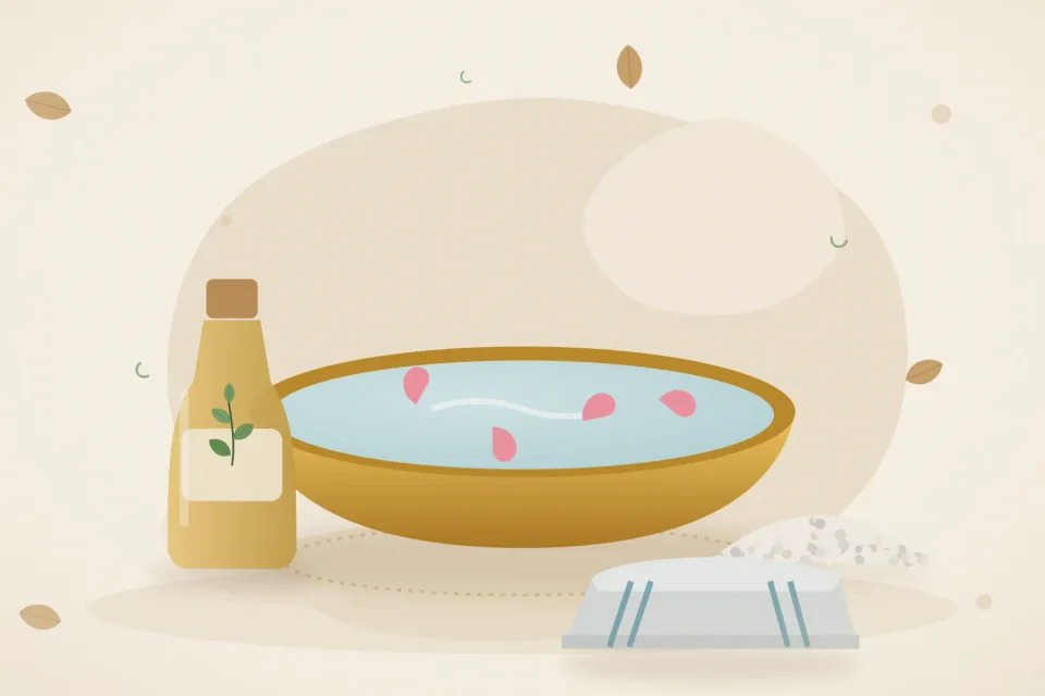 A brass basin of warm water with petals, a bottle of sesame oil, a small bowl of salt and a folded towel on a sandy beige background