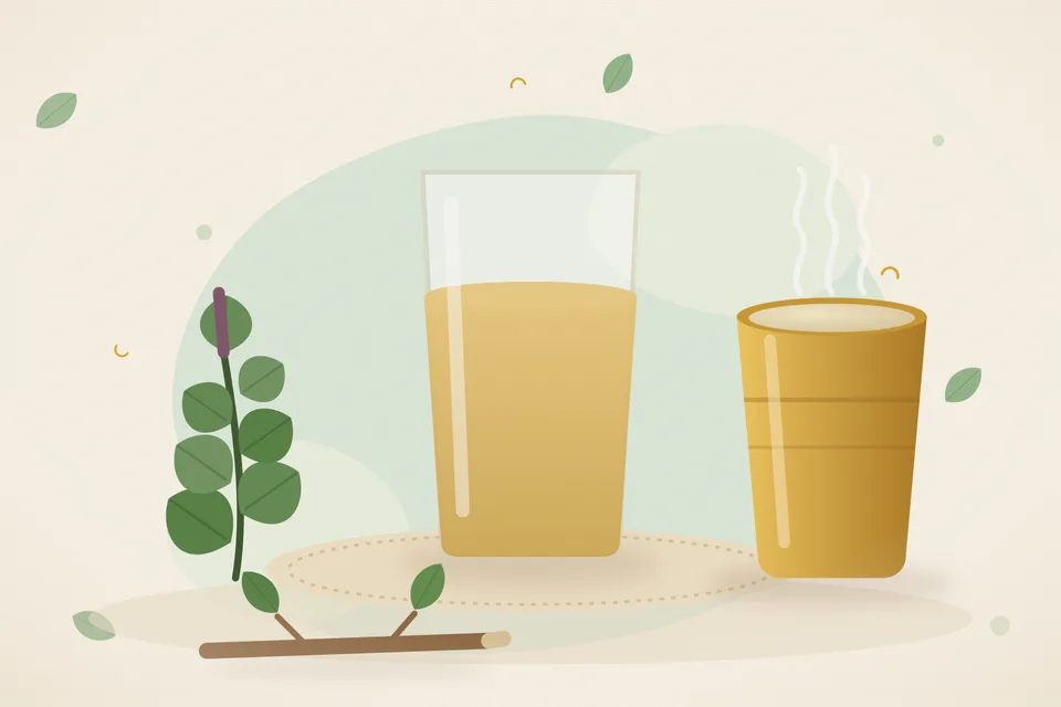 A small steel pot of tulsi leaves in water on a kitchen stove, with a cup of cooled rinse beside a wooden board.